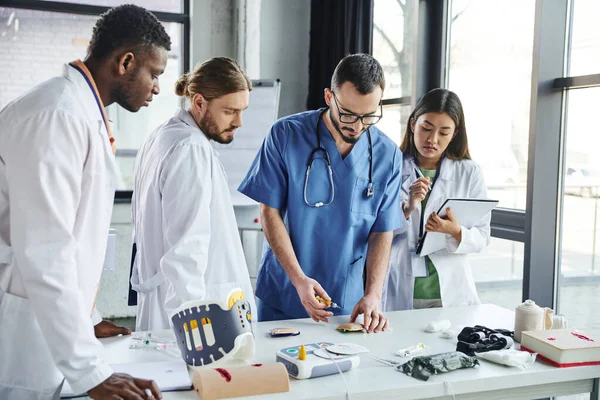 Diverse group of multiethnic students in white coats looking at healthcare instructor making injection in training pad near medical equipment, skills development concept — Stock Photo
