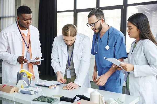 Young student in white coat practicing with injection training pad near professional paramedic and multiethnic team with notebooks near medical equipment, skills development concept — Stock Photo