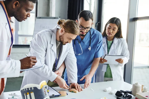Young student making injection in training pad near medical equipment, healthcare worker and multiethnic team in white coats during first aid seminar, skills development concept — Stock Photo