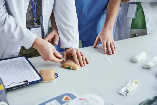 Partial view of student in white coat doing injection in training pad near clipboard, bandages and healthcare worker during medical first aid seminar, life-saving skills development concept — Stock Photo