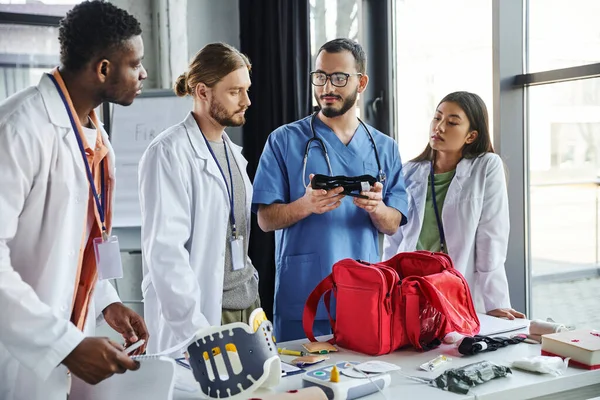 Healthcare worker in uniform showing compressive tourniquet to diverse group of interracial students near medical equipment in training room, life-saving skills and bleeding prevention concept — Stock Photo