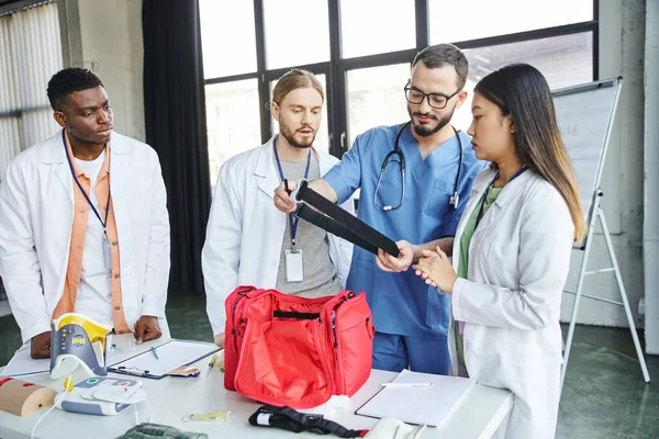 First aid seminar, paramedic in uniform and eyeglasses showing compressive tourniquet to interracial team near first aid kit and medical equipment, life-saving skills and bleeding prevention concept — Stock Photo