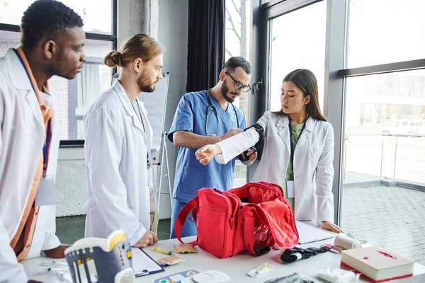 Trabajador sanitario en anteojos y uniforme aplicando torniquete compresivo en brazo de mujer asiática cerca de estudiantes multiétnicos, botiquín de primeros auxilios y equipo médico, concepto de prevención de hemorragias — Stock Photo