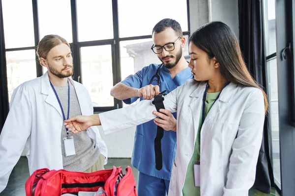 First aid hands-on learning, young paramedic in eyeglasses and uniform applying compressive tourniquet on arm of asian student in white coat, life-saving skills and bleeding prevention concept — Stock Photo