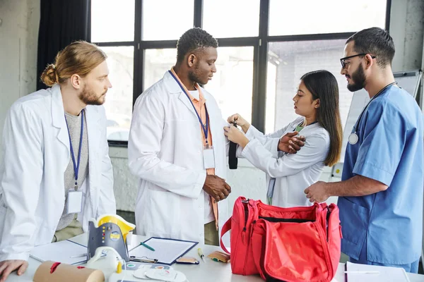 Asian woman in white coat applying compressive tourniquet on arm of african american student near paramedic, first aid kit and medical equipment, life-saving skills and bleeding prevention concept — Stock Photo