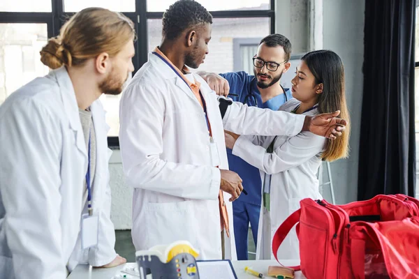 Healthcare worker helping asian student applying compressive tourniquet on arm of african american man near first aid kit and medical equipment, life-saving skills and bleeding prevention concept — Stock Photo