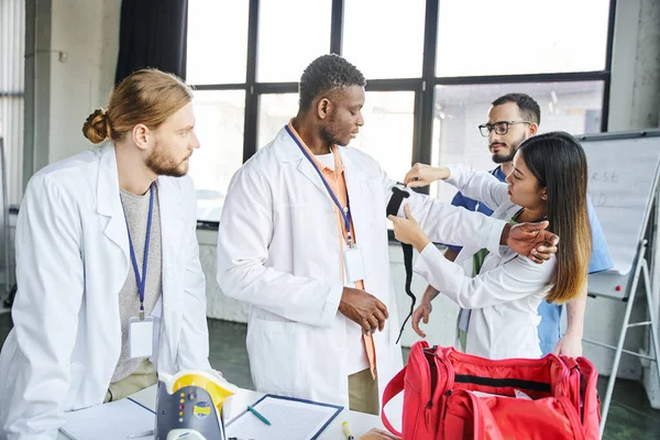 Asian woman in white coat applying compressive tourniquet on arm of african american student near instructor, medical equipment and first aid kit, life-saving skills and bleeding prevention concept — Stock Photo