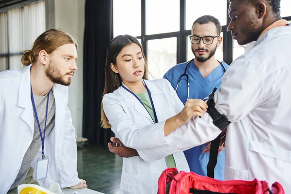 Paramedic in eyeglasses looking at young asian woman applying compressive tourniquet on arm of african american student during first aid seminar in training room, bleeding prevention concept — Stock Photo