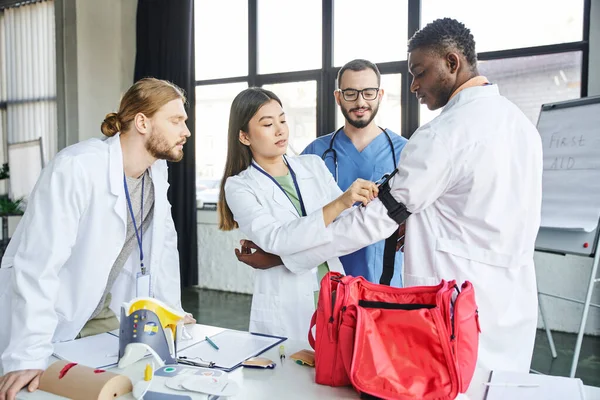 Young asian woman applying compressive tourniquet on arm of african american man near first aid kit, medical equipment and doctor with interracial students, bleeding prevention concept — Stock Photo