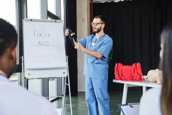 Doctor in blue uniform and eyeglasses standing with compressive tourniquet near flip chart with first aid lettering and multiethnic students on blurred foreground, life-saving skills concept — Stock Photo