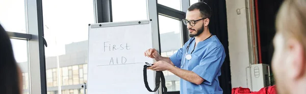 Young healthcare worker in eyeglasses and blue uniform showing compressive tourniquet near flip chart with first aid lettering, emergency preparedness and life-saving skills concept, banner — Stock Photo