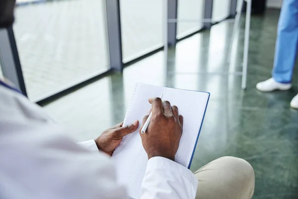 Cropped view of african american student in white coat sitting with notebook and pen near medical instructor on blurred background, emergency preparedness and life-saving skills development concept — Stock Photo