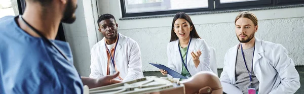 Young asian woman asking question to medical instructor with CPR manikin during first aid seminar with interracial students, acquiring and practicing life-saving skills concept, banner — Stock Photo