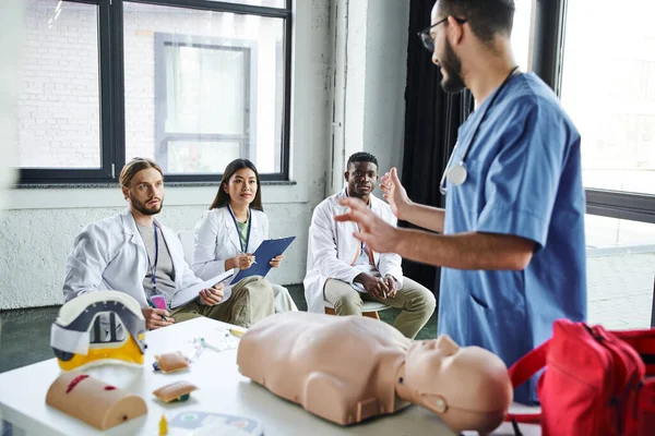 Travailleur de la santé en uniforme bleu parler à des étudiants multiethniques en blouse blanche près du mannequin CPR et de l'équipement médical dans la salle d'entraînement, l'acquisition et la pratique des compétences de sauvetage concept — Photo de stock