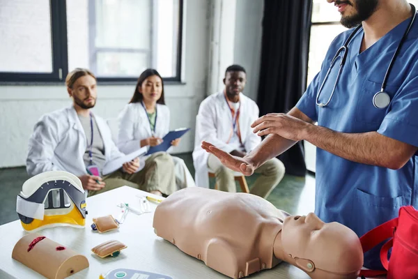 Healthcare worker showing life-saving techniques on CPR manikin near medical equipment and diverse group of interracial students on blurred background, acquiring life-saving skills concept — Stock Photo
