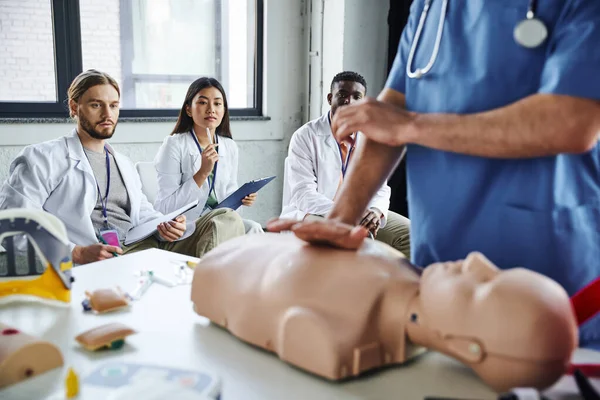 Young multiethnic students in white coats looking at paramedic showing life-saving techniques on CPR manikin on blurred foreground, acquiring and practicing life-saving skills concept — Stock Photo