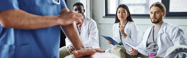 Diverse group of interracial students in white coats looking at medical instructor showing life-saving skills on blurred foreground, acquiring and practicing life-saving skills concept, banner — Stock Photo