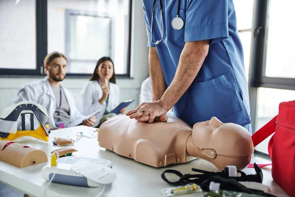 Professional paramedic in blue uniform doing chest compressions on CPR manikin near medical equipment and young multiethnic students on blurred background, practicing life-saving skills concept — Stock Photo