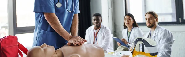 Professional paramedic doing chest compressions on CPR manikin near multiethnic students in white coats during first aid seminar, acquiring and practicing life-saving skills concept, banner — Stock Photo