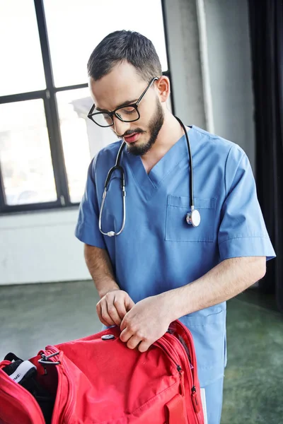 Молодой и бородатый медицинский работник в очках и синей форме расстегивая красную сумку первой помощи во время подготовки к медицинскому семинару, приобретая концепцию жизненно важных навыков — стоковое фото