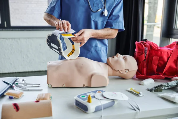 Partial view of healthcare worker in blue uniform holding neck brace above CPR manikin, first aid kit, defibrillator and medical devices in training room, life-saving skills development concept — Stock Photo