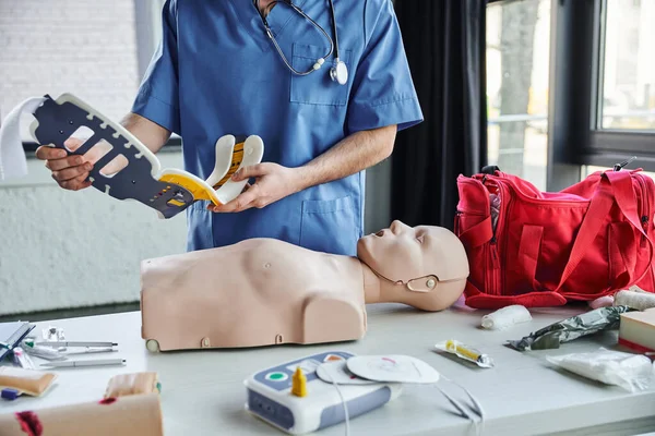 Partial view of healthcare worker in blue uniform holding neck brace near CPR manikin, defibrillator, first aid kit and medical equipment, life-saving skills development concept — Stock Photo