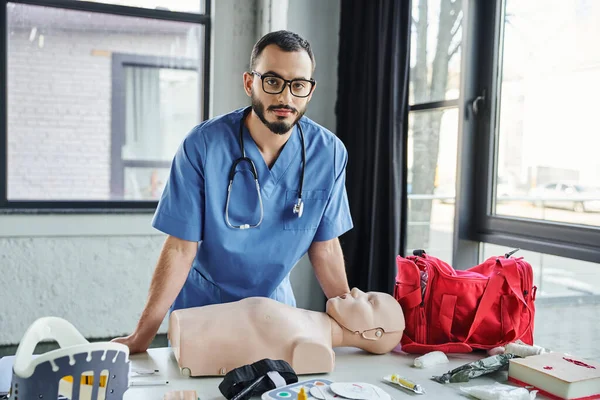 Ambulancier paramédical barbu avec lunettes et uniforme bleu regardant la caméra près du mannequin CPR, sac de premiers soins rouge, défibrillateur et orthèse de cou dans la salle d'entraînement, concept de développement des compétences vitales — Photo de stock
