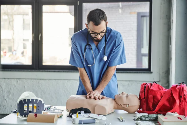 Young paramedic in blue uniform and eyeglasses practicing chest compressions on CPR manikin near defibrillator and first aid kit during medical seminar, life-saving skills development concept — Stock Photo