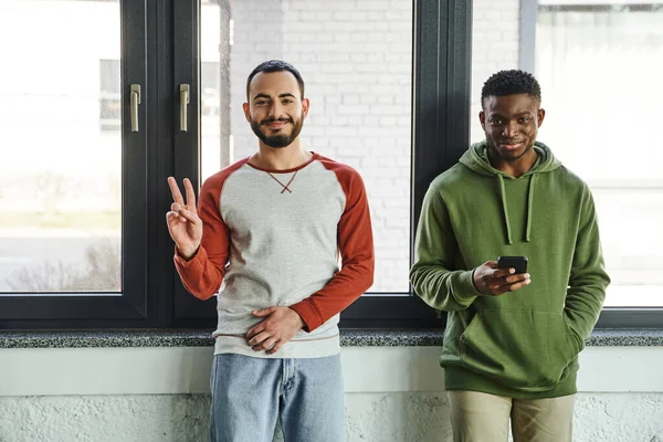Young interracial business colleagues in stylish casual clothes looking at camera in modern office, cheerful bearded man showing victory sign near african american man networking on mobile phone — Stock Photo