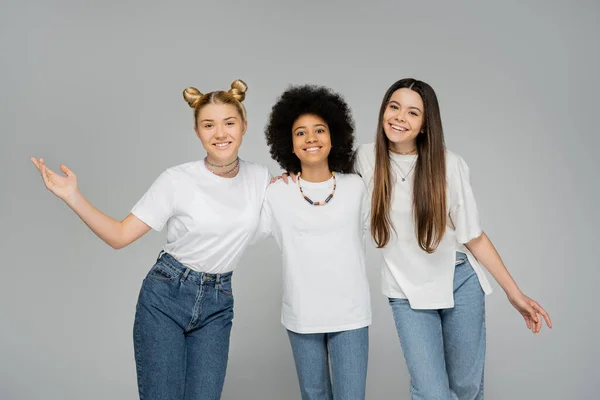 Cheerful and multiethnic teenage girlfriends in white t-shirts hugging each other and looking at camera while standing isolated on grey, teenage friends having fun together — Stock Photo