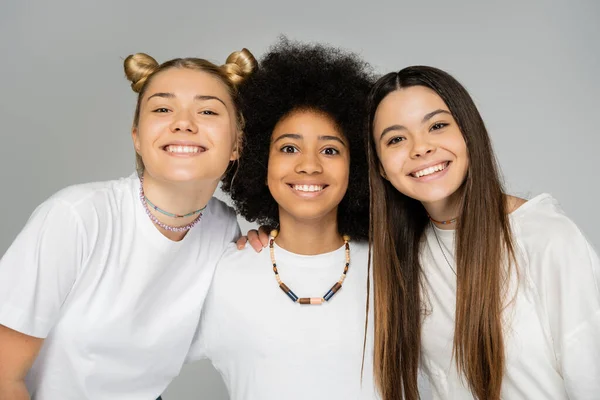 Portrait of positive interracial girlfriends in white t-shirts hugging african american friend and looking at camera together isolated on grey, teenage friends having fun together — Stock Photo