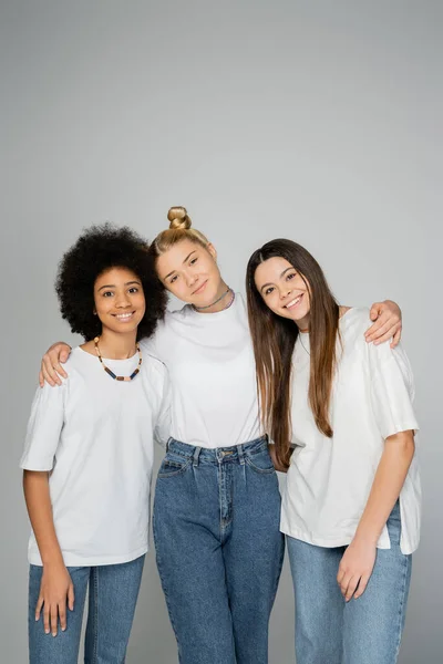 Stylish and cheerful interracial teenage girls in white t-shirts and blue jeans hugging and looking at camera while standing isolated on grey, teenage friends having fun together — Stock Photo
