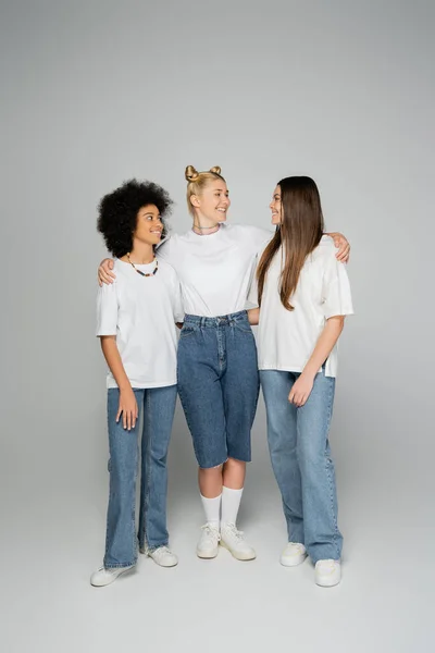 Полная длина улыбающаяся блондинка-подросток обнимает многоэтнических девушек в белых футболках и синих джинсах, стоя и разговаривая на сером фоне, друзья-подростки веселятся вместе — стоковое фото