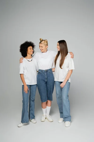 Full length of cheerful blonde teen girl hugging positive multiethnic girlfriends in white t-shirts and stylish blue jeans while standing on grey background, teenage friends having fun together — Stock Photo