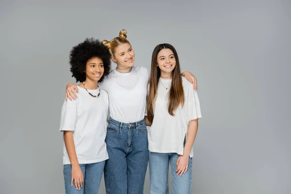 Smiling blonde teenage girl in white t-shirt and blue jeans hugging multiethnic friends and looking away while standing isolated on grey, teenage friends having fun together — Stock Photo