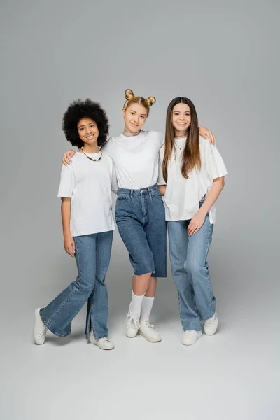 Full length of smiling and stylish multiethnic teenage girlfriends hugging and looking at camera while standing and posing together on grey background, multiethnic teen models concept — Stock Photo