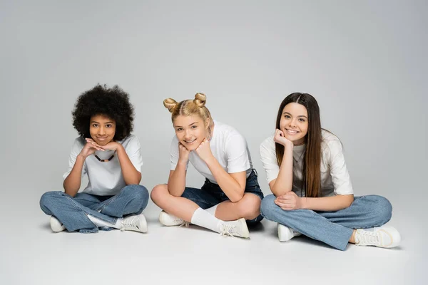 Ganzer Länge lächelnde Teenager-Mädchen in weißem T-Shirt und stylischer blauer Jeans, die in die Kamera schauen, während sie zusammen sitzen und Zeit auf grauem Hintergrund verbringen, Konzept multiethnischer Teenie-Modelle — Stockfoto