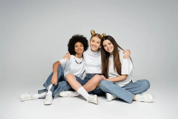 Joyful and multiethnic teenage girls in stylish white t-shirts and blue jeans hugging and looking at camera while sitting together on grey background, multiethnic teen models concept — Stock Photo