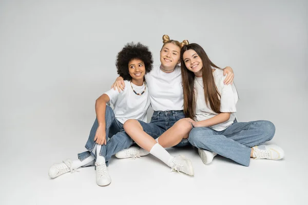 Comprimento total de menina adolescente loira abraçando alegres namoradas multiétnicas em elegantes camisetas brancas e jeans enquanto olha para o fundo cinza, conceito de modelos adolescentes multiétnicos — Fotografia de Stock