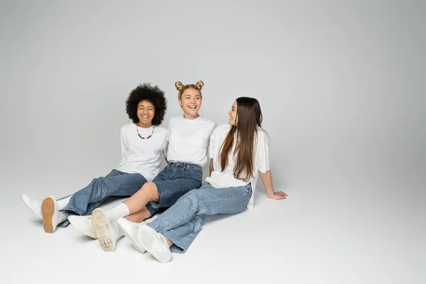 Ganzer Länge positiver und multiethnischer Teenager-Mädchen in weißen T-Shirts und blauen Jeans, die sich unterhalten, während sie sitzen und Zeit miteinander auf grauem Hintergrund verbringen, Konzept multiethnischer Teenie-Modelle — Stockfoto