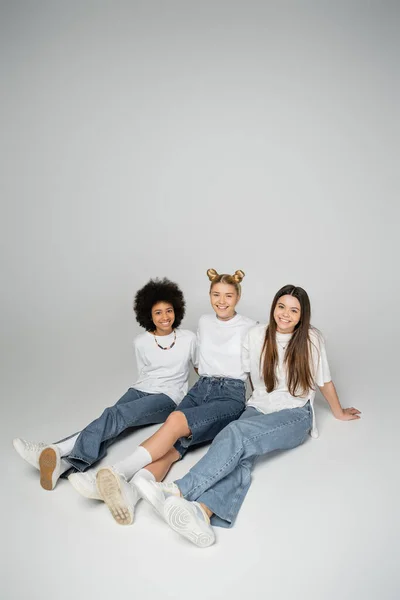 Весёлые и многоэтнические девушки-подростки в модных джинсах и белых футболках, смотрящие в камеру, сидя и позируя на сером фоне, концепция мультиэтнических моделей подростков — стоковое фото