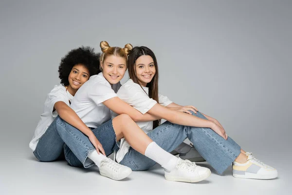 Повна довжина усміхнених і багатоетнічних дівчат-підлітків у білих футболках і блакитних джинсах, які дивляться на камеру, сидячи і позуючи на сірому фоні, концепція багатоетнічних моделей підлітків — стокове фото