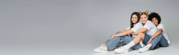 Pleine longueur de copines multiethniques adolescentes positives en t-shirts et jeans décontractés regardant la caméra tout en étant assis sur fond gris, concept de modèles adolescentes multiethniques, bannière avec espace de copie — Photo de stock