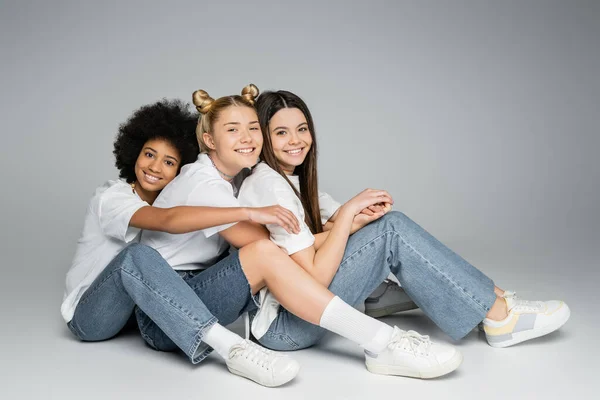 Full length of positive and multiethnic teenage friends in casual white t-shirts and jeans looking at camera and sitting on grey background, multiethnic teen models concept, friendship and bonding — Stock Photo