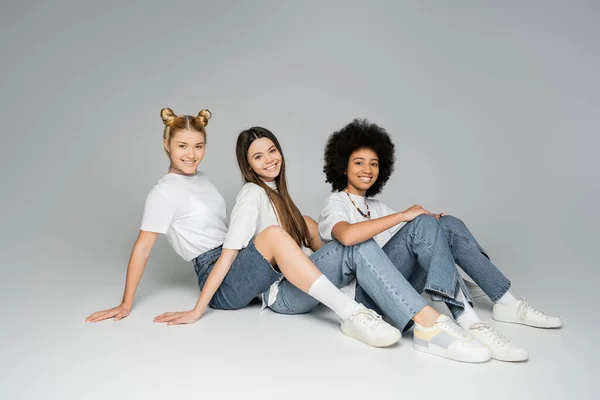 Полная длина положительных и подростковых многонациональных девушек в повседневных футболках и джинсах, смотрящих в камеру, сидя вместе на сером фоне, концепция мультиэтнических моделей подростков — стоковое фото