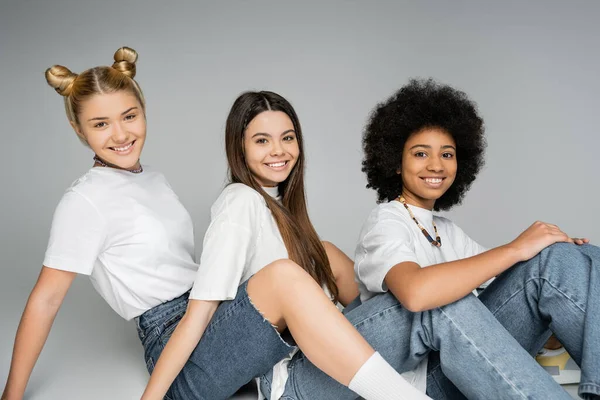 Радісні підліткові багатоетнічні подружки в білих футболках і блакитних джинсах дивляться на камеру, сидячи і позуючи разом на сірому фоні, концепція багатоетнічних моделей підлітків, дружба і зв'язок — стокове фото