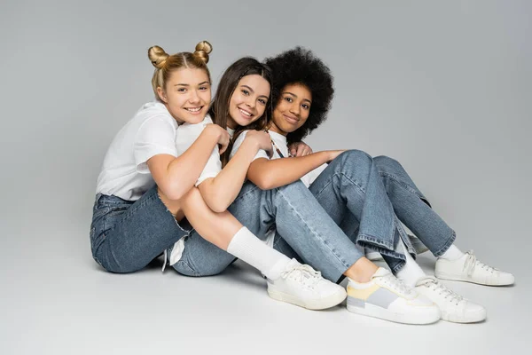 Full length of teen and cheerful multiethnic girlfriends in white t-shirts and jeans hugging and posing together on grey background, multiethnic teen models concept, friendship and bonding — Stock Photo