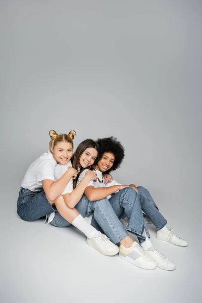 Pleine longueur d'amis positifs et adolescents multiethniques en t-shirts blancs décontractés, jeans et baskets regardant la caméra et assis sur fond gris, concept de modèles adolescents multiethniques — Photo de stock
