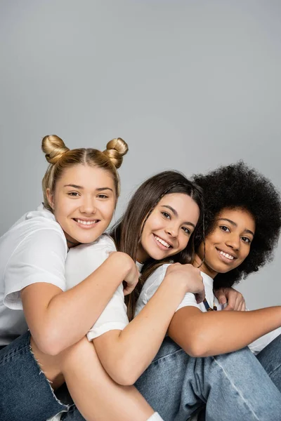 Portrait of positive multiethnic teenage girls in white t-shirts and jeans hugging and posing next to each other isolated on grey, multiethnic teen models concept, friendship and bonding — Stock Photo