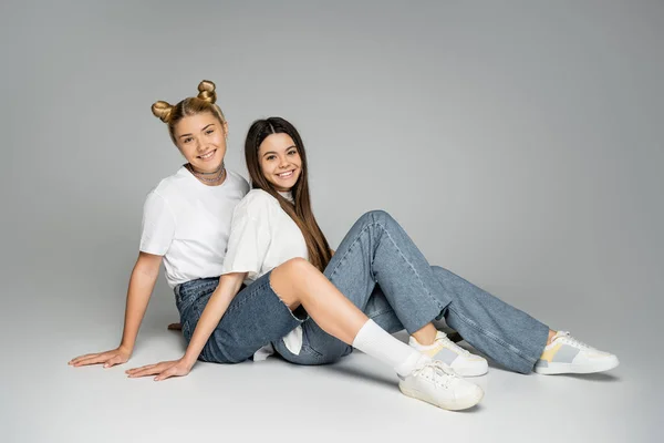 Meninas adolescentes loiras e morenas sorridentes em camisetas brancas, jeans e tênis olhando para a câmera enquanto se sentam juntas em fundo cinza, conceito de modelos adolescentes, amizade e vínculo — Fotografia de Stock